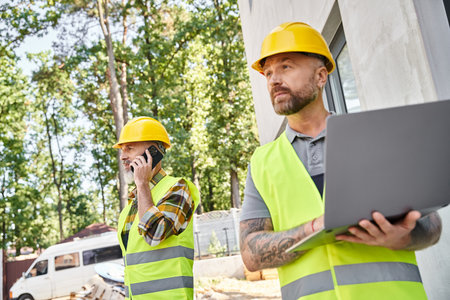 appealing builder with tattoos working on laptop while his colleague talking by phone during work