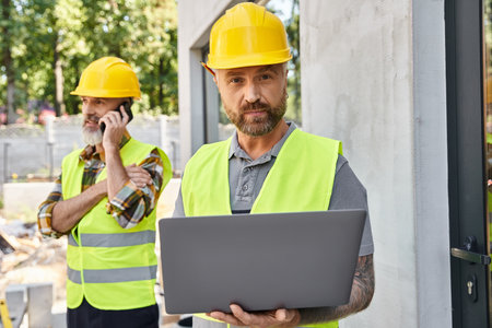 appealing builder with tattoos working on laptop while his colleague talking by phone during workの写真素材