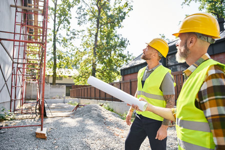 two good looking cottage builders in safety helmets with blueprints working near scaffolding