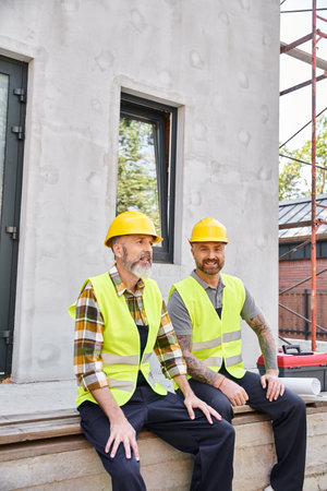 cheerful hardworking cottage builders in safety helmets sitting on porch and looking at camera