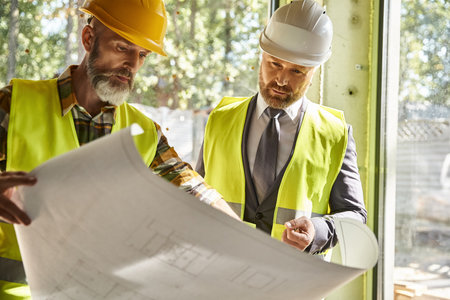 handsome bearded businessman and cottage builder with safety gear working with blueprint on site