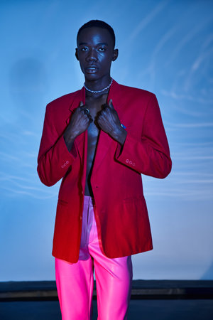 alluring stylish african american man in pink pants and red blazer posing on blue watery backdrop