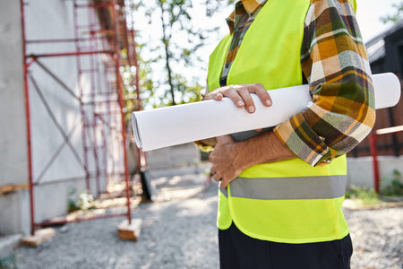 cropped view of dedicated cottage builder with blueprint in hand posing next to scaffolding