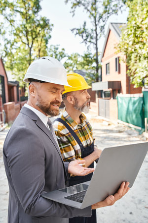 joyous handsome architect and builder in overalls and suit working with laptop on construction site