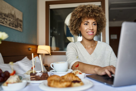 joyful african american woman using her laptop near breakfast in hotel, room service and convenienceの写真素材