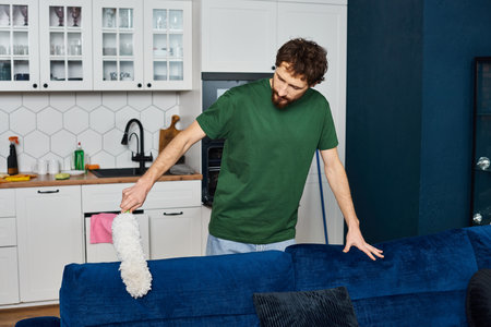 good looking man in comfy casual outfit cleaning sofa with duster with kitchen on background
