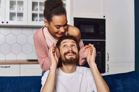 handsome man looking at his african american girlfriend after she surprised him and closed eyes