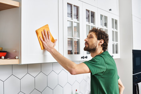 good looking man in casual t shirt using rag to clean kitchen cupboards at home, spring cleaningの写真素材