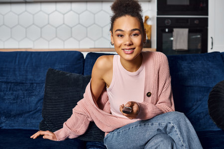 beautiful african american jolly woman sitting on sofa with remote control and looking at camera