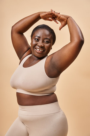 smiling plus size woman in beige underwear posing with raised hands, body positive and self esteemの写真素材