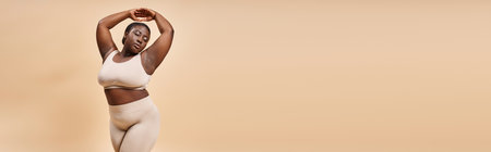 plus size african american woman in underwear posing with raised hands on beige backdrop, banner