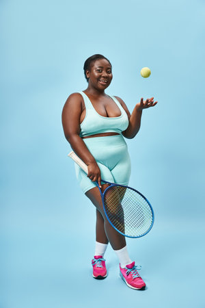 cheerful plus size african american woman holding tennis racket and throwing ball on blue backdropの写真素材