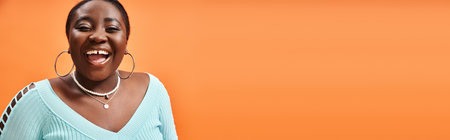 portrait of cheerful plus size african american woman in blue long sleeve laughing on orange, banner