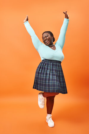 cheerful plus size african american model in plaid skirt and mint color jumper posing on orange