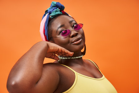 Fashionable and plus size african american woman in headscarf and trendy sunglasses on orangeの写真素材
