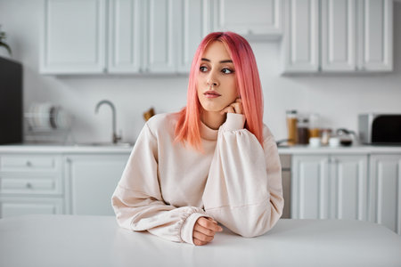 good looking jolly woman with pink hair in casual attire sitting and looking away in kitchenの写真素材