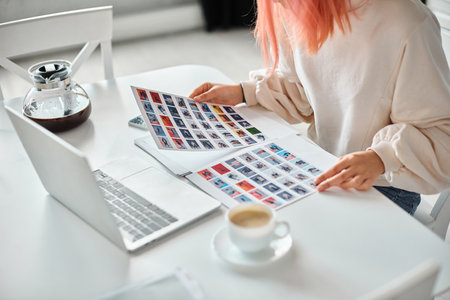cropped view of young woman with pink hair holding papers with images next to her laptop at homeの写真素材