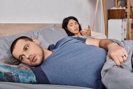 young frustrated tattooed man looking away while lying down on bed near asian wife, family conflictの写真素材