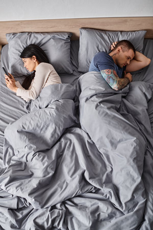 young tattooed man near asian wife looking at his smartphone in bedroom, misunderstanding, top view