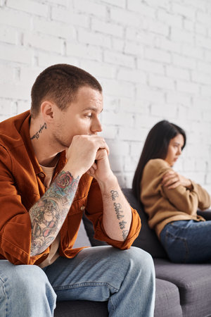 frustrated tattooed man looking at offended asian wife sitting on couch at home, divorce conceptの写真素材