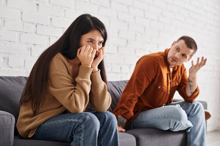 discouraged man talking to offended asian wife sitting on couch in living room, divorce conceptの写真素材