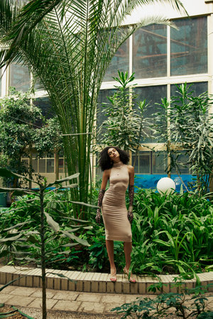 full length of african american woman in dress and animal print gloves posing among plants in gardenの写真素材