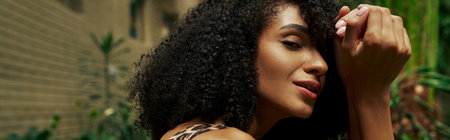 beautiful african american curly woman in chic attire smiling and posing around green plants, bannerの写真素材