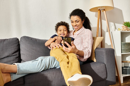 cheerful african american mother hugging her little son and looking at smartphone together with himの写真素材