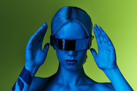 futuristic blonde woman with sci fi robotic sunglasses posing in blue lights on green backdropの写真素材