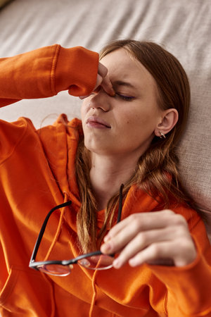 top view of tired teen girl in orange hoodie leaning on sofa at home, melancholy and solitudeの写真素材