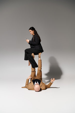 young businesswoman with smartphone balancing on feet of man in suit on grey, couple of acrobats