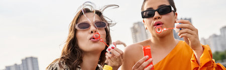 beautiful jolly women in vibrant outfits with sunglasses blowing soap bubbles on rooftop, banner