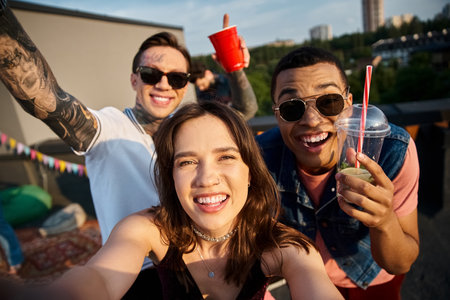 cheerful multicultural friends holding cocktails in hands and having fun at rooftop party