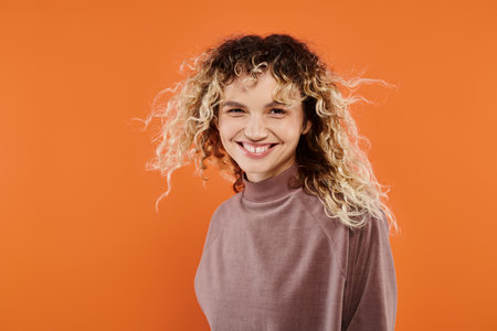 cheerful woman with wavy hair in mocha color turtleneck looking at camera on radiant orange backdrop
