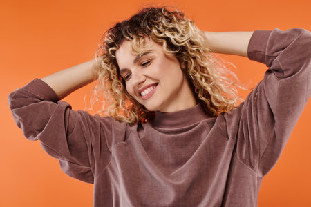 portrait of attractive smiling woman with wavy hair in brown turtleneck on orange background