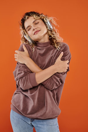 dreamy woman with wavy hair listening music in wireless headphones and hugging herself on orange