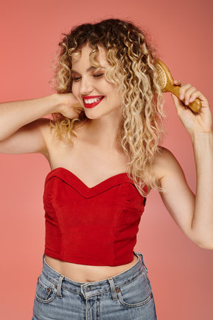 smiling curly and attractive woman in red top brushing wavy hair on pastel pink studio backdrop