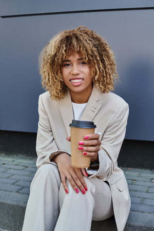 smiling african american woman in suit sitting on parapet with coffeee to go and looking at camera