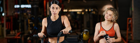 energetic sporty woman using modern cross trainer in gym next to her athletic female coach, banner