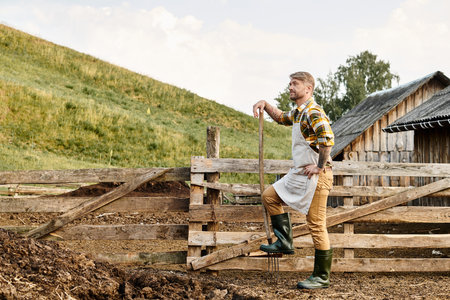 hard working attractive farmer with beard and tattoos using pitchfork while working with manure