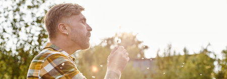 cheerful appealing man with tattoos blowing on dandelion while chilling on farm in sunlight, banner