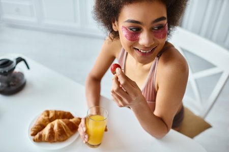 happy african american woman eating holding strawberry and glass of orange juice in kitchenの写真素材
