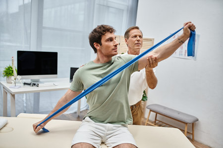 attractive man stretching his muscles with resistance band near his mature doctor, rehabilitation