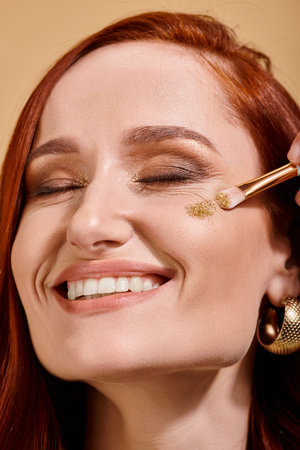 Radiant and redhead woman applying sparkling glitter on cheek with makeup brush on beige backdrop