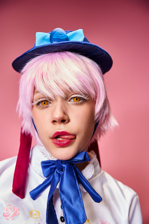 alluring young female cosplayer sticking out her tongue and looking at camera on pink backdrop