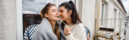 smiling lesbian woman feeding her girlfriend while having breakfast on balcony, lgbt couple bannerの写真素材