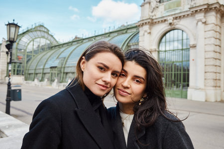 cheerful lesbian couple in coats looking at camera near Palmenhaus in Vienna on backgroundの写真素材