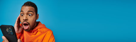 banner of surprised african american man scrolling social media in smartphone on blue backgroundの写真素材