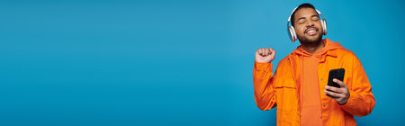 banner of smiling african american man in orange outfit and headphones dancing with smartphone