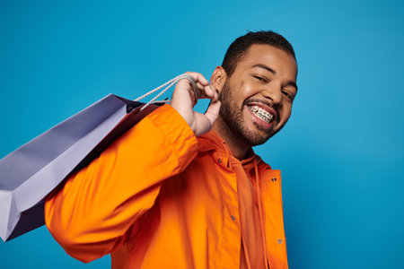 smiling african american man in orange outfit with shopping bag over shoulder on blue backgroundの写真素材